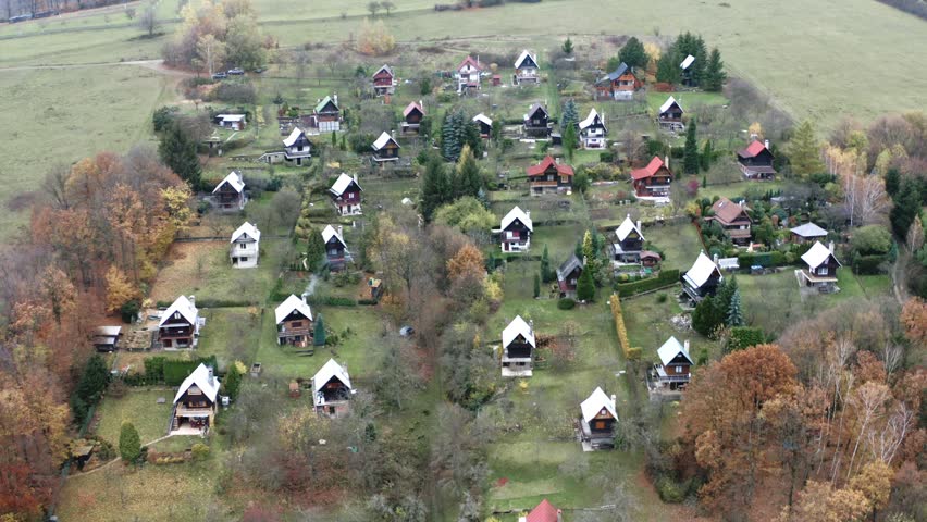 Aerial drone shot capturing small wooden houses nestled within autumn landscape in rural area, cinematic footage featuring village settlement among trees, peaceful countryside scenery.