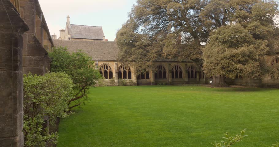 Lush green quadrangle lawn is framed by cloister arches and mature trees at New College Oxford on a calm day.