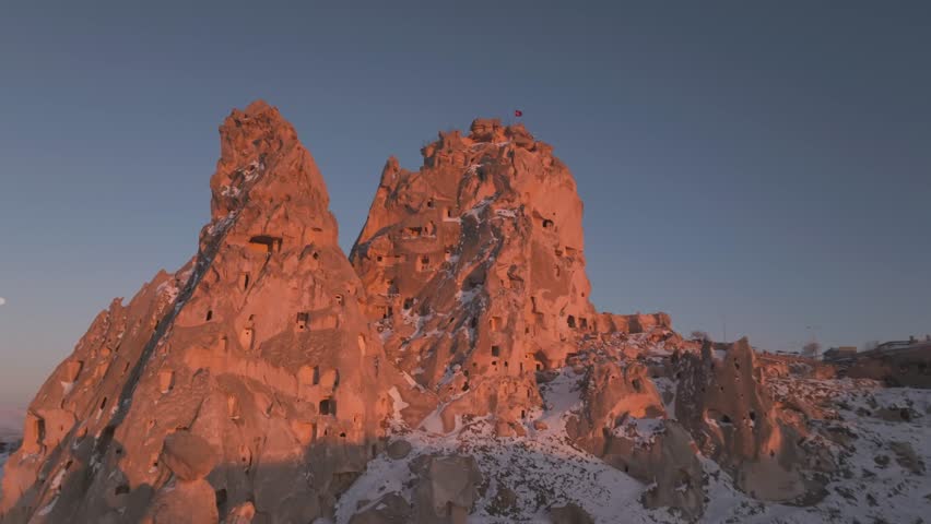 Drone aerial of Uchisar Castle in Cappadocia, Turkey during golden hour. Historic rock fortress overlooking snowy valleys, cave dwellings and the unique volcanic landscape of Cappadocia.