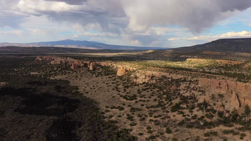 4K UHD drone video capturing majestic sandstone cliffs stretching across a vast southwest landscape. Dark volcanic lava flows frame the foreground while rain clouds gather over distant mountains.