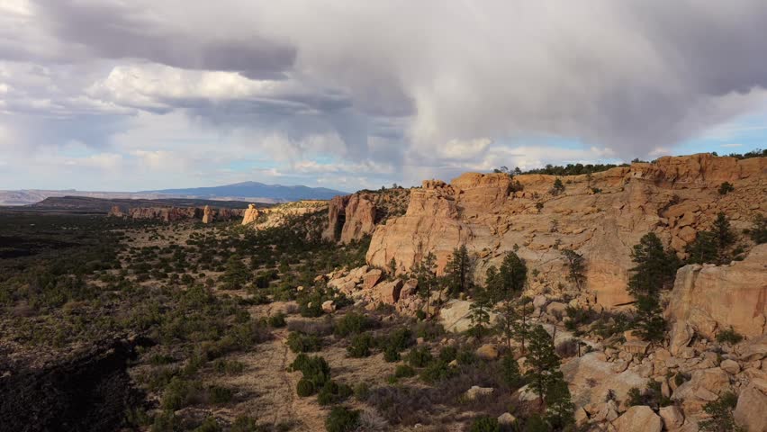 4K UHD drone flight revealing a pristine desert wilderness where golden sandstone cliffs rise above ancient black lava fields. Heavy rain showers sweep across the distant mountain backdrop.