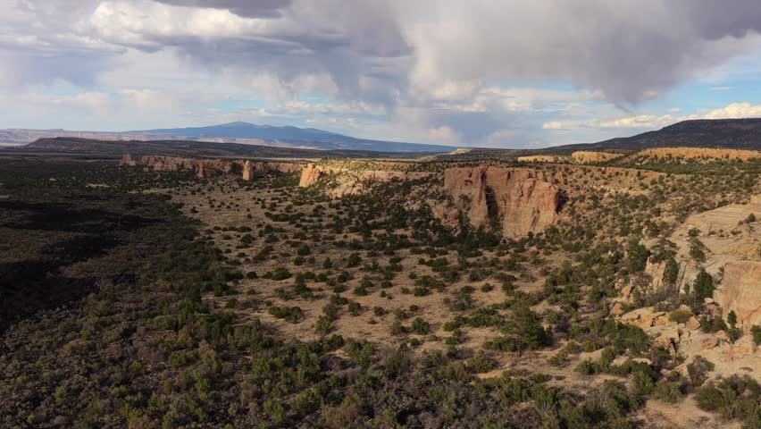4K UHD drone flight showcasing a pristine southwest wilderness where towering sandstone cliffs meet ancient black lava beds. Distant rain showers fall over the rugged mountain horizon at dusk.
