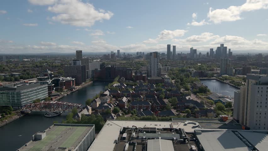 Aerial footage of Salford Quays waterfront with contemporary buildings and bridge, showcasing modern Manchester. Drone moving forward over the buildings with the river and bridgeon the left side.
