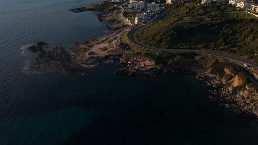 Curving coastal road leads toward the seaside town of Alghero as a drone glides above rocky shoreline at golden hour.