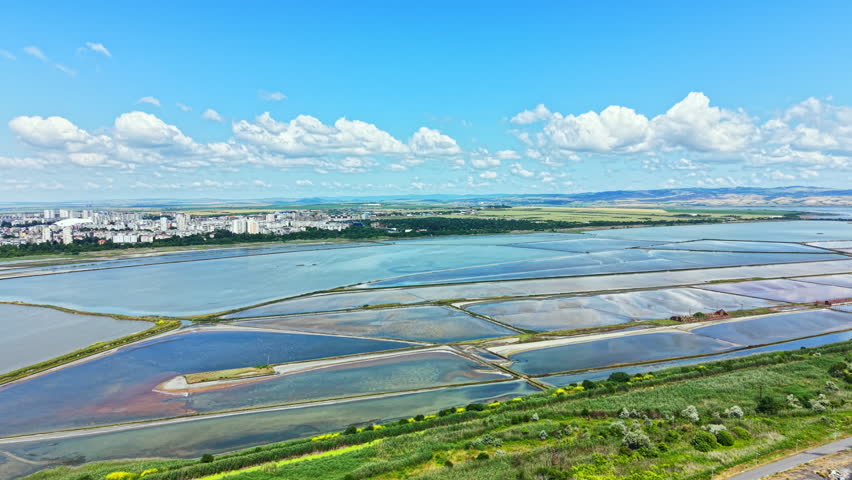 The scene shows salt pans in Burgas with varying water levels and colors. The sky is blue and filled with clouds. The city and hills can be seen in the background.