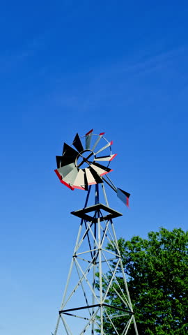 A windmill spins slowly in the breeze under a clear blue sky. Trees are nearby, offering shade on a bright sunny day. The scene shows a typical rural setting with few clouds in the sky.