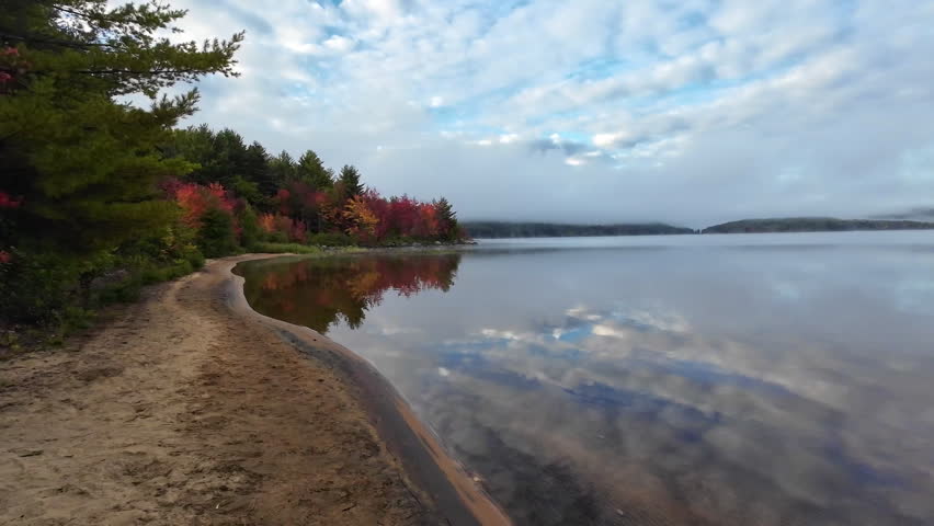 Still water reflecting clouds beside colorful autumn trees along the shoreline of Grand Lake in Achray, Algonquin Provincial Park, Ontario, Canada, with low clouds and light fog over the landscape.