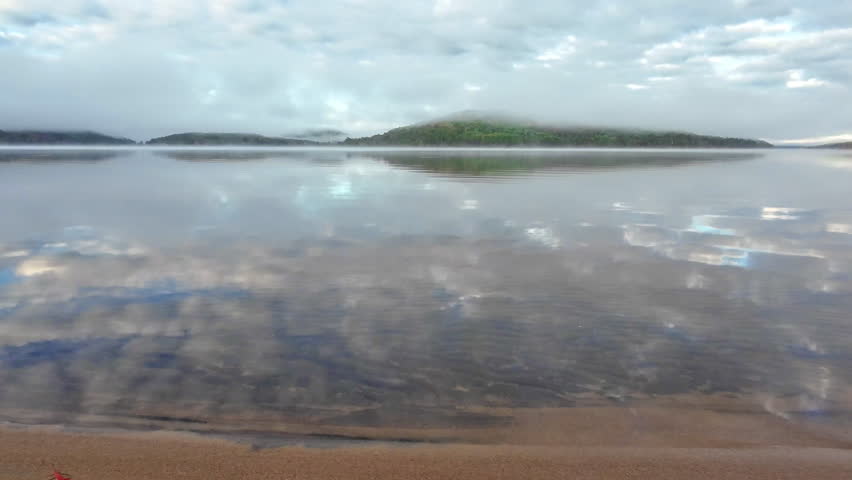 Calm water reflecting clouds above a sandy shoreline on Grand Lake in Achray, Algonquin Provincial Park, Ontario, Canada, with low clouds and fog over the distant forested hills.