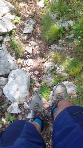 First-person perspective video capturing the lower legs of an adventurous male hiker sitting down to rest before continuing to walk along a narrow, rugged mountain path on a bright, sunny summer afternoon. The hiker is wearing dark blue athletic shorts, grey trekking shoes with orange laces, and vibrant blue socks. The camera angle looks directly downward from the hiker