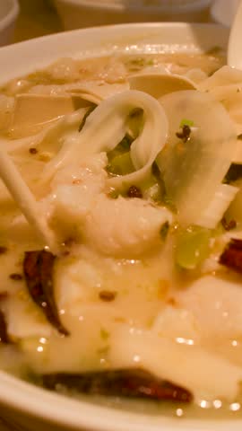 Close-up shot of chopsticks lifting fish and noodles from a spicy ceramic bowl of soup.