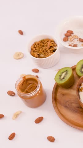 Hands scoop peanut butter into a yogurt bowl with berries and nuts on white background.