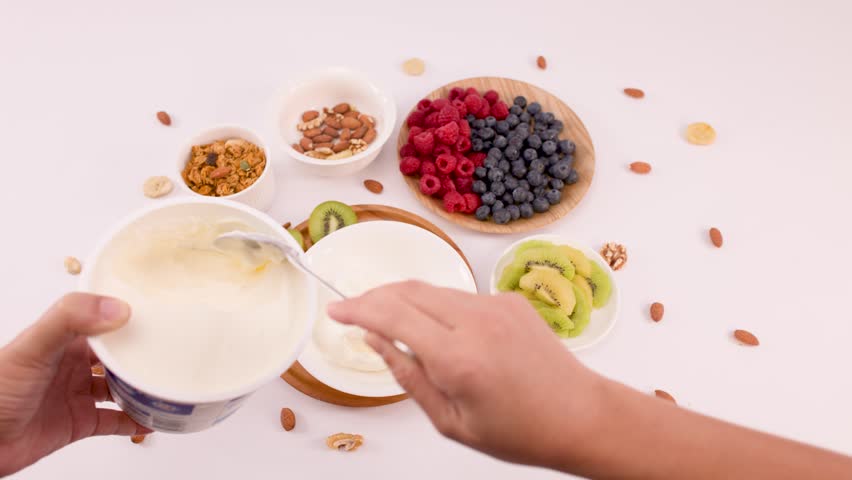 Overhead shot of hands scooping white yogurt into a bowl surrounded by fresh fruit.