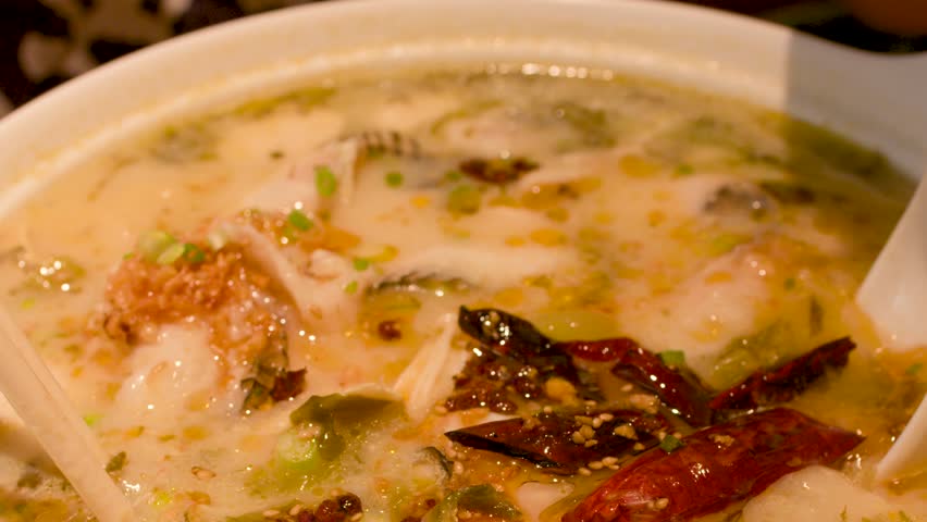 Close-up shot of chopsticks stirring spicy fish noodle soup in a ceramic bowl with chili.