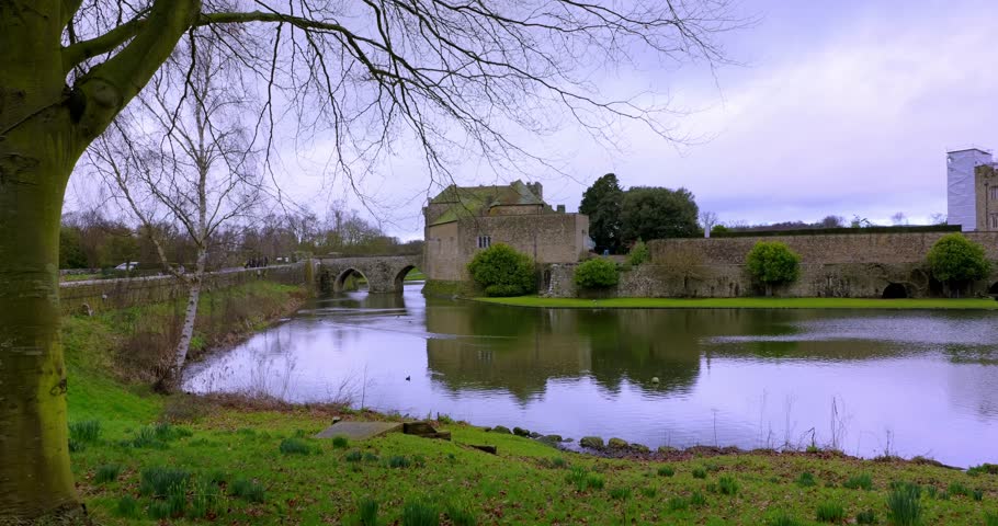 Wide landscape view featuring Leeds Castle, its moat and stone bridge surrounded by tranquil grounds.