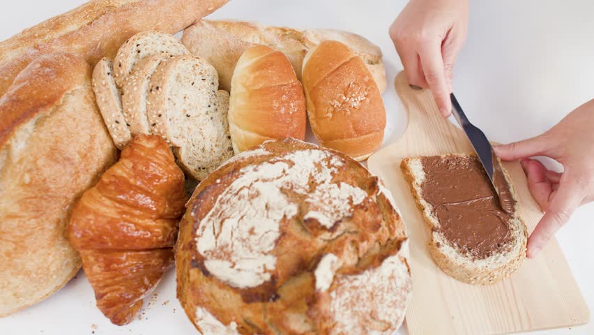 Hands spread chocolate cream on bread beside sourdough, croissants, and baguettes on white background.