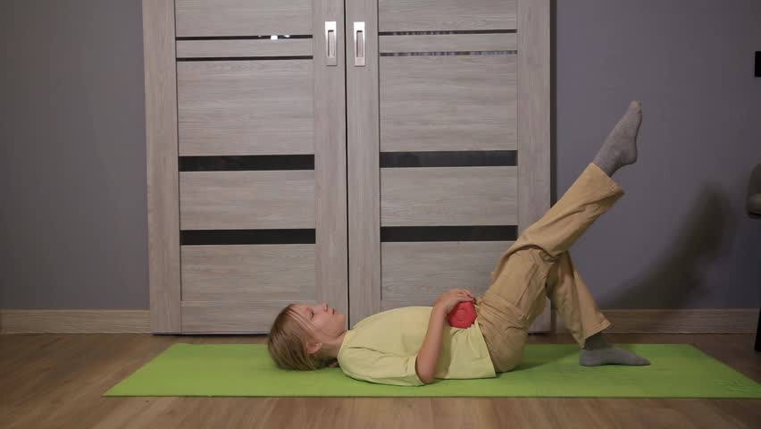 Side view of young girl lying on green yoga mat, performing single-leg bridge with one leg elevated, small red dumbbell on hips, demonstrating home workout and muscle development.