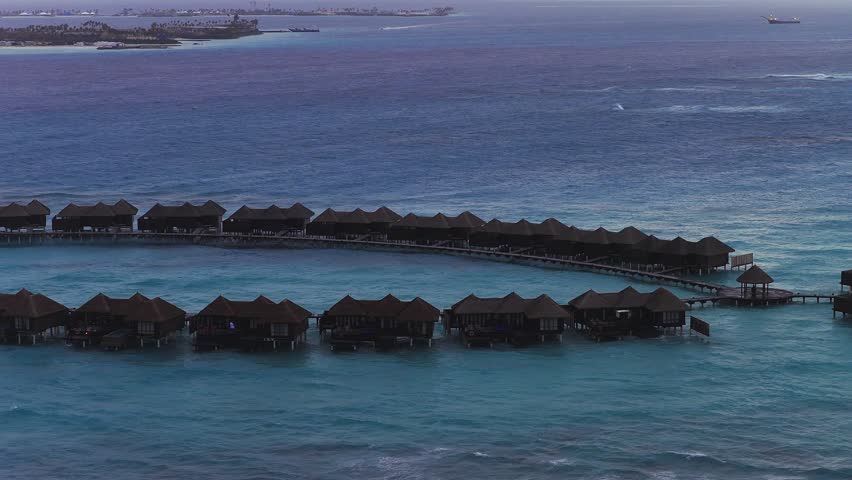 Aerial view of a Maldives resort shows arcs of over water villas, wooden walkways, a gazebo, a bright turquoise lagoon, and the deeper Indian Ocean with a distant cargo ship.