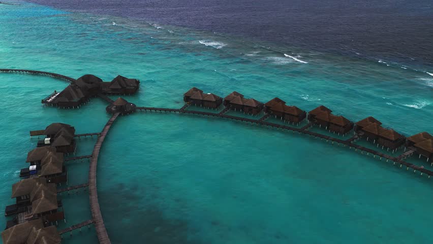Aerial view shows a Maldives resort of water villas on stilts, curved wooden walkways, a circular pavilion, and a palm island, as a drone sweeps over a turquoise lagoon.