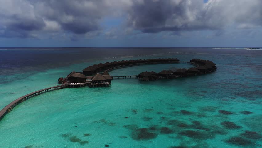 Aerial daylight view of a Maldives overwater resort, a semicircle of thatched villas on stilts and walkways above a turquoise lagoon, as the drone moves outward under clouds.