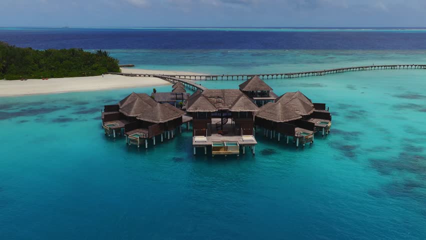 Aerial drone sweep reveals thatched overwater villas with plunge pools in a Maldives lagoon, a curving wooden jetty to a white sand island, and reef bands at the horizon.
