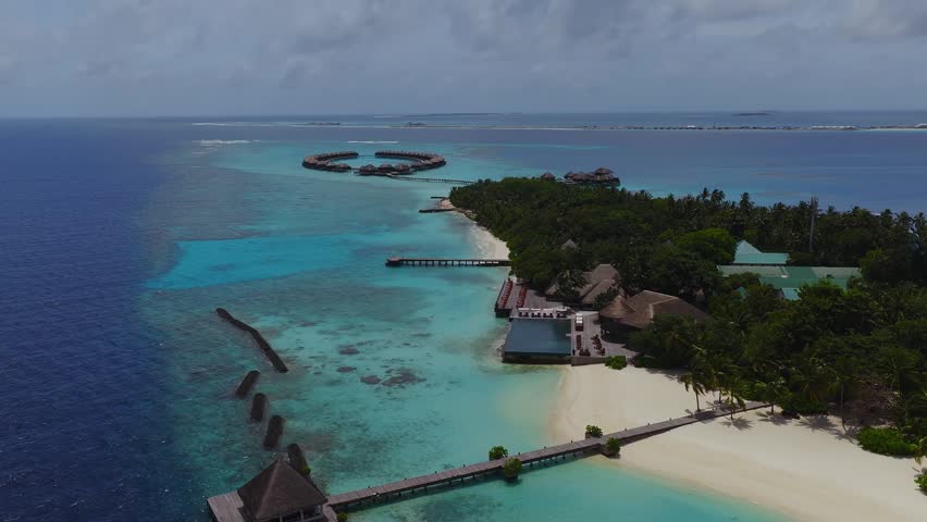 Aerial view of a Maldives island resort with ring shaped overwater villas, long piers, an infinity pool, white sand, palm groves, and a turquoise lagoon next to deep ocean.