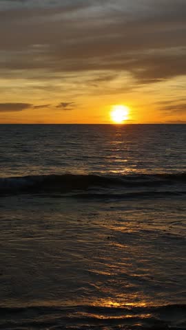 Beautiful seascape featuring the sun setting over the ocean horizon, casting warm golden light across gentle waves beneath a dramatic sky filled with colorful clouds. This tranquil coastal scene captures the beauty of nature, travel, relaxation, and the peaceful atmosphere of twilight by the sea.