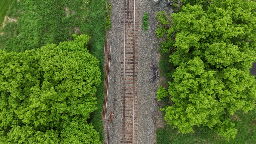 Railway tracks in southern Manitoba. Drone looks straight down and moves forward above rail line crossing a small creek with green grass, trees, rock ballast, and cloud reflections on a summer day.