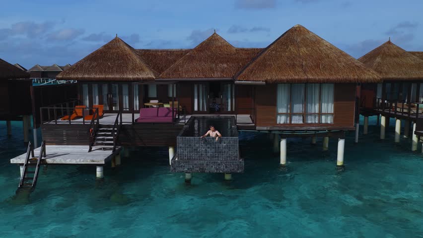 Aerial view of a Maldives resort with thatched villas on stilts, private pools, curved walkway, turquoise shallows, coral patterns, and the Indian Ocean beyond.