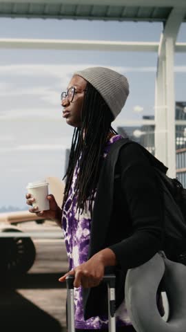 Vertical frame footage: Woman in busy airport terminal traveling to destination with coffee cup in hand, while young traveler carrying trolley in airport hallway, holding drink container.