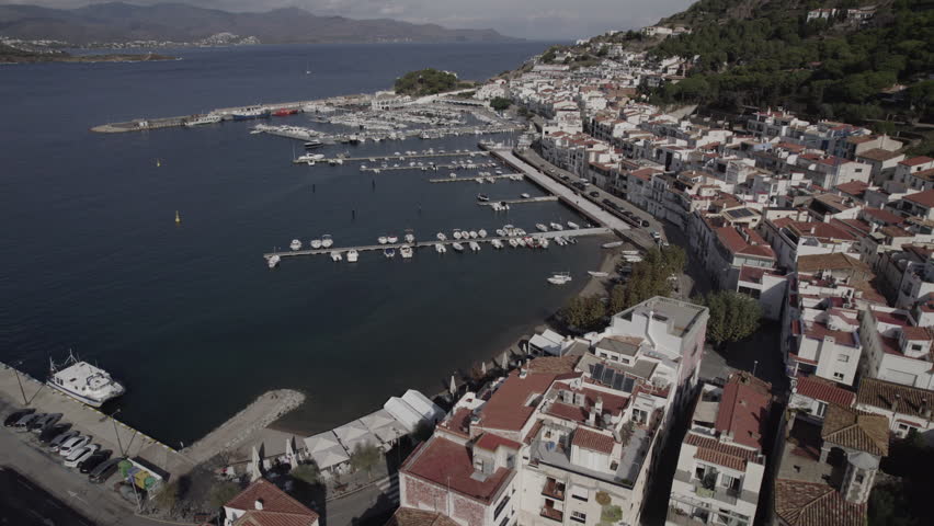 Aerial drone footage flying forward at higher altitude towards the coastal town of Port de la Selva, Costa Brava, Catalonia, Spain, offering a wide elevated view of the Mediterranean seaside village and surrounding coastline.