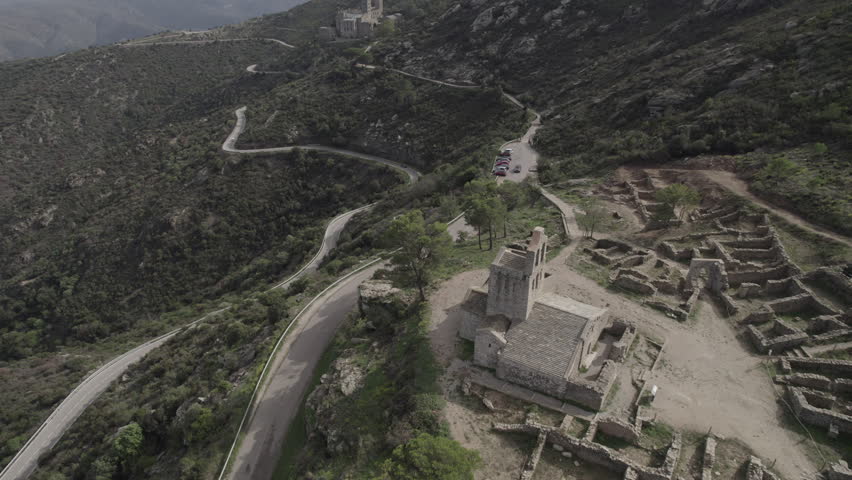 Aerial drone footage from high altitude following the pathway that connects the church ruins to the Monastery of Sant Pere de Rodes, Costa Brava, Catalonia, Spain, showing the full route within the mountain landscape.