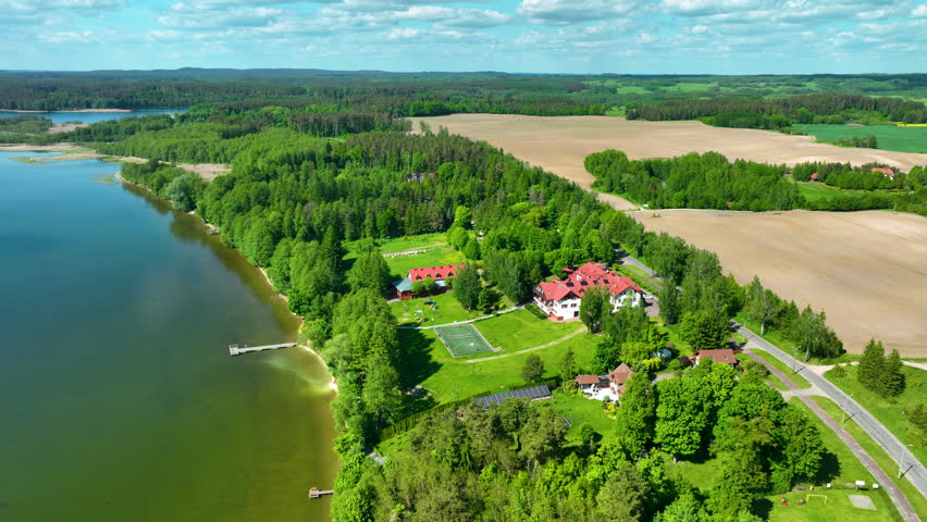 Cinematic high angle shot of a lush lakeside resort surrounded by green forests and agricultural fields in the Masurian Lake District, Poland.