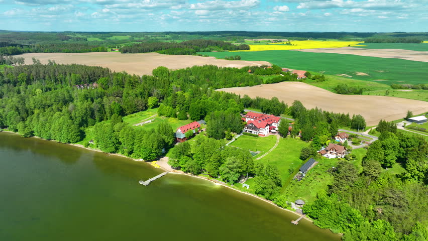 Cinematic aerial shot of a lakeside resort hotel in the Masuria region of Poland, featuring lush forests, farmland, and clear blue water for travel and wellness concepts.