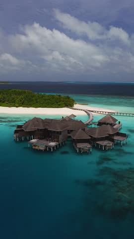 Aerial view shows thatched overwater villas with private pools, glass platforms, and a curving wooden jetty in the Maldives as the camera glides and descends.