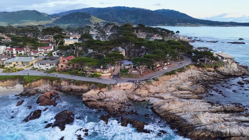 Aerial drone view of a rocky California coastline with cypress trees, luxury homes, and ocean waves, resembling Carmel-by-the-Sea.