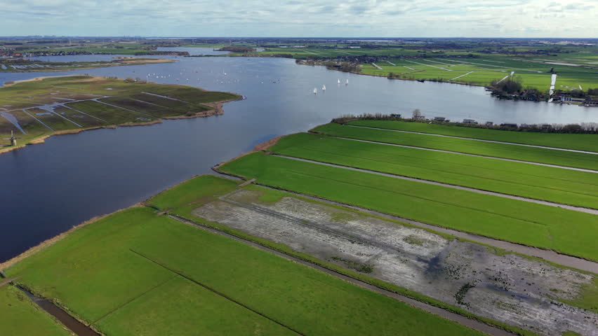 A high-angle aerial drone photograph capturing the traditional polder landscape of the Dutch countryside. On the far left, a historic windmill stands at the edge of the wetlands. The composition showcases a vast network of drainage canals and geometric ditches cutting through vibrant green agricultural pastures. A wide river channel or lake opens up in the background, where dozens of white sailboats and leisure yachts are scattered across the water. The opposite shore is lined with small village houses, clusters of trees, and infinite lowlands extending to the horizon under a cloudy spring sky. This panoramic daytime view beautifully represents European water management, maritime heritage, and seasonal travel destinations.