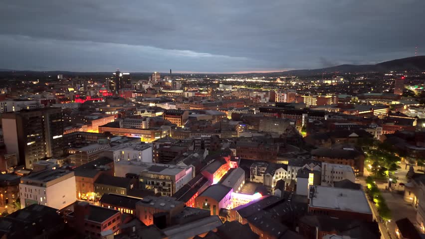 Slow drone flight over illuminated urban landscape during dusk showing historic architecture and modern streets. Cinematic footage captures night life and glowing city lights in UK.