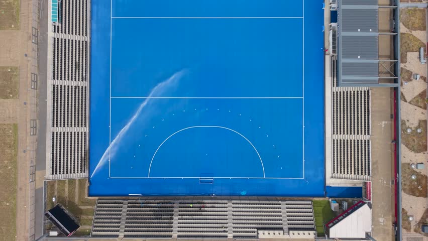 Blue hockey pitch from above as the watering cannon shuts off, leaving wet turf and spray marks at Lee Valley Hockey and Tennis Centre in Stratford, London, UK.