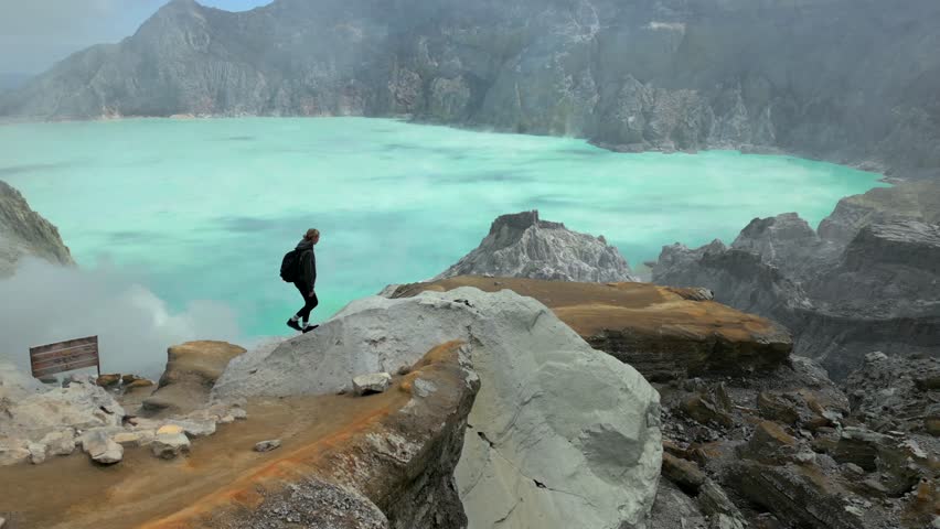 Young woman hiking along the rim of Kawah Ijen crater overlooking the turquoise acidic lake in East Java, Indonesia. Dramatic volcanic landscape with rugged cliffs and sulfur mist.