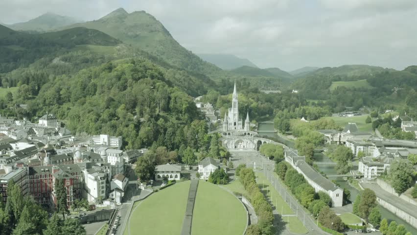 Aerial forward view of the Sanctuary of Notre-Dame de Lourdes in Hautes-Pyrénées, France, featuring the renowned pilgrimage church, surrounding hills, religious buildings, and sacred sanctuary complex, capturing spiritual heritage and architectural beauty.
