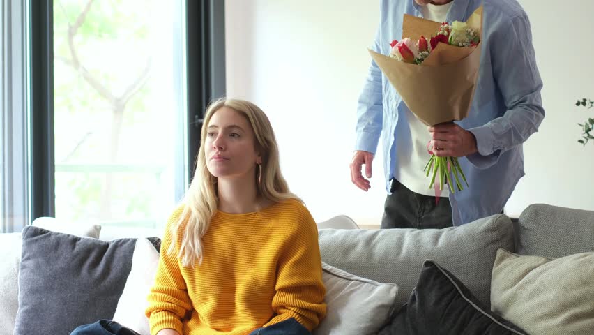 A loving man surprises his girlfriend with a beautiful bouquet of fresh flowers, bringing a look of joyful surprise and happy emotions to her face in their comfortable home setting.
