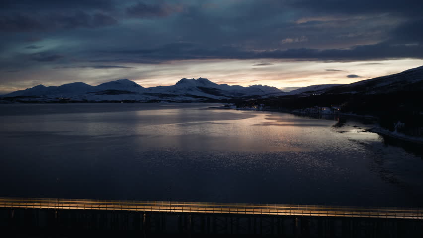 Snowy Arctic mountains beyond a calm Norwegian fjord at blue hour beneath dramatic clouds, drone establishing