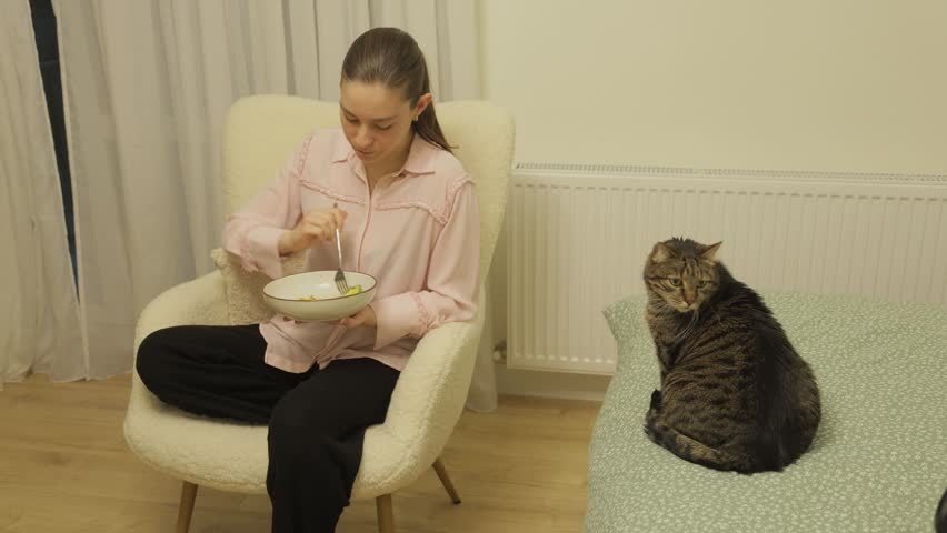 Young woman eating a healthy meal while her cute tabby cat sits on the bed nearby, attentively watching her every move with curiosity