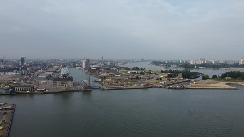 Drone flyover above Antwerp’s Port House, revealing the dock basin, the Scheldt River, and the city skyline beneath bright overcast skies.