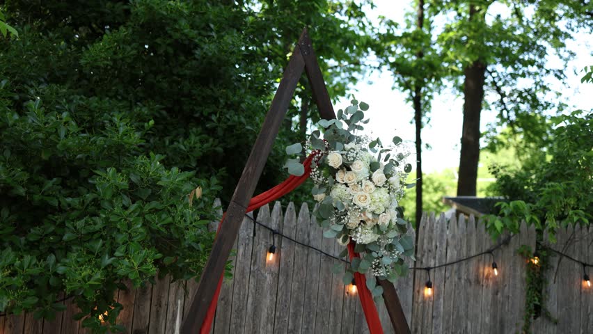 Rustic outdoor wedding arch with white flowers, green leaves and red fabric beside a wooden fence and string lights in a leafy garden setting