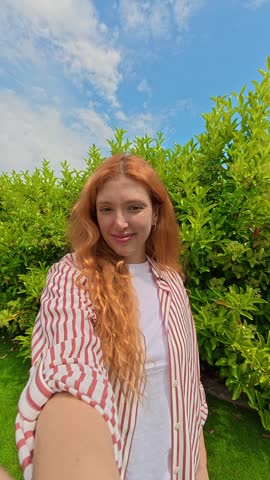 A happy redhead woman with a bright smile enjoys a beautiful day outdoors, posing for a cheerful selfie against a backdrop of green nature and blue sky.