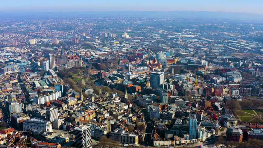 Aerial drone view of Bristol city centre, England, showing mixed urban architecture, church spires, modern office towers, construction cranes, and surrounding residential areas in winter.