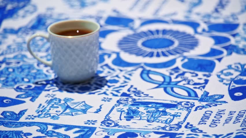 A hand placing a plate of pastel de nata tarts served beside a coffee cup on a blue and white tablecloth of traditional Portuguese azulejo tile motifs. Static shot, selective focus. Iconic dessert, local cuisine, food tourism, unique experience. Translation: best things in life are not things.