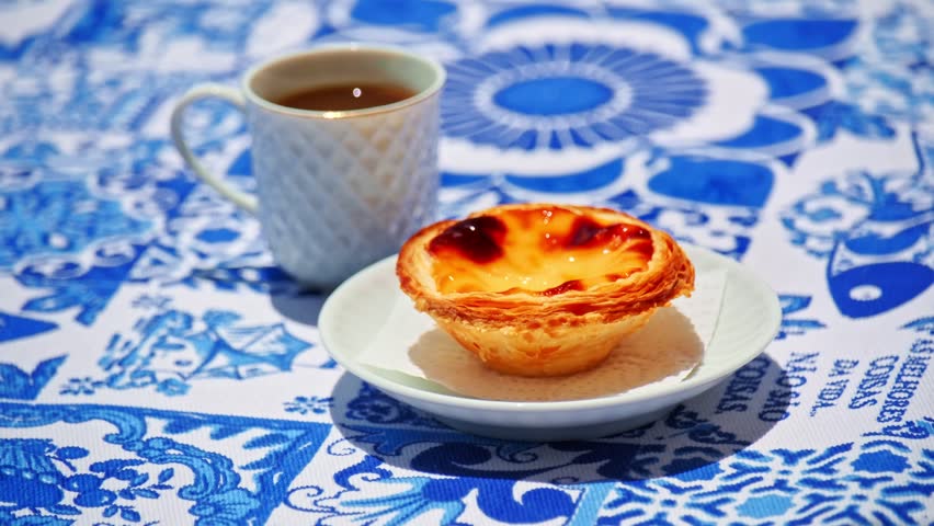 Traditional Portuguese pastry known as pastel de nata custard tart touched and taken by hand on white plate beside porcelain coffee cup on blue and white tablecloth with azulejo motifs. Famous heritage pastry, local specialty, food experience. Translation: best things in life are not things.