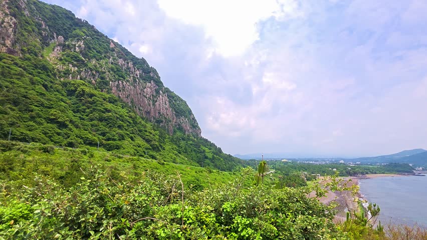 Pan left from hiking trail with red railing revealing towering Sanbangsan mountain lava dome with rugged cliffs and green shrubs on sunny summer day in Jeju, South Korea